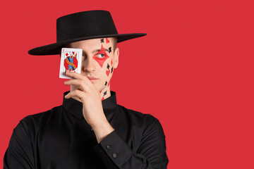 Young man with drawn card suits on his face against red background, closeup
