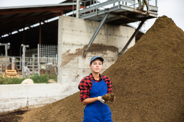 Female farmer with shovel poses against pile of dried and crushed cow dung in the backyard of a farm