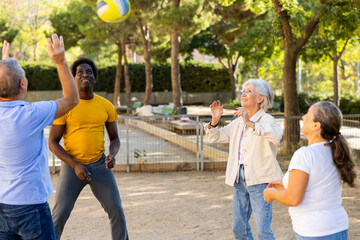 Positive multiracial friends of different ages throwing up a volleyball ball in a circle on the sand in the park