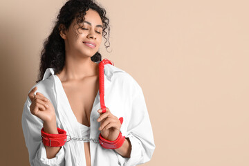 Young African-American woman with whip and handcuffs from sex shop on beige background