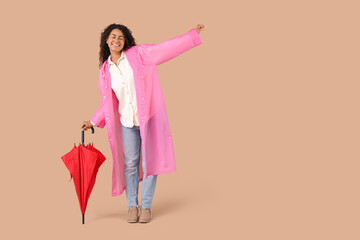 Happy young African-American woman in raincoat with umbrella on beige background