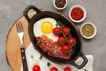 Pan with fried egg, bacon, tomatoes and bowls of spices on wooden stand on grey background. Closeup