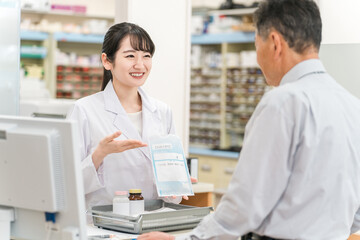 薬局のレジで薬の説明をする薬剤師の女性と高齢者男性
