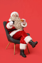 Santa Claus with tasty burger and soda sitting in armchair on red background