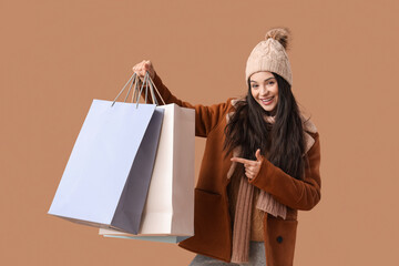 Beautiful young woman in warm clothes pointing at shopping bags on brown background