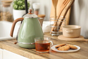 Modern green electric kettle with glass cup of hot tea and sweet cookies on counter in kitchen