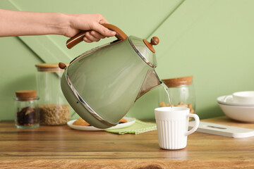 Woman pouring water from modern green electric kettle into cup on counter in kitchen