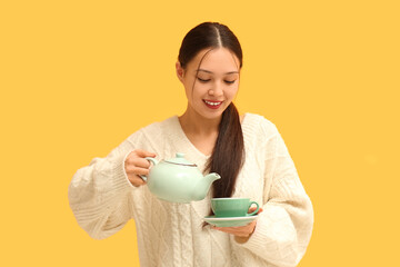 Beautiful young Asian woman with cup of hot tea and teapot on yellow background