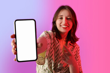 Fototapeta premium Young woman pointing at blank mobile phone on color background