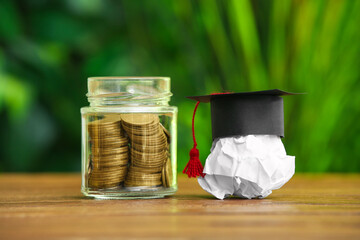 Crumpled paper with graduation hat and jar of coins on table outdoors. Student loan concept