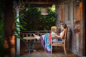 Young woman reading on open-air veranda of wooden bungalow with tropical garden view. Girl relaxing...