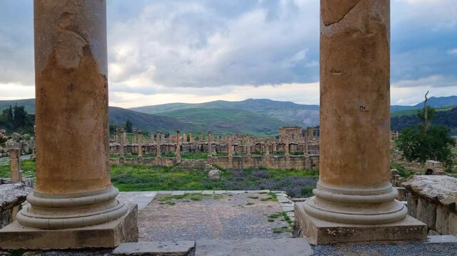 Revealing shot filmed between the entrance columns of the Septimius Severus Temple in Djemila, Algeria, showcasing the ancient settlement and the green hillside landscape on a cloudy day