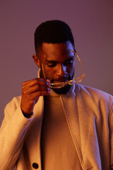 Stylish young Black man adjusting his glasses while wearing a cozy beige coat, exuding a thoughtful expression against a softly lit purple background