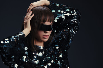 Woman in sequinned dress poses against a black background, wearing sunglasses on her head, hands...