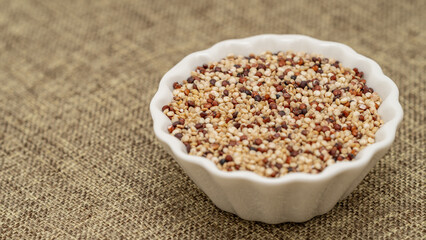 Mixed quinoa grains in a white ceramic bowl placed on a textured fabric background, featuring red, white, and black seeds. Ideal for healthy eating and food concepts.