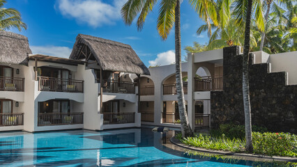 Fototapeta premium Swimming pool at the resort. Cottages with thatched roofs and terraces around the perimeter. Palm trees, green grass in a flower bed. Shadows on clear aquamarine water. Blue sky, clouds. Mauritius.
