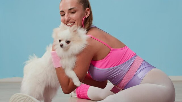 Tilt up slowmo portrait of happy Caucasian female athlete in pink and purple 80s sportswear hugging fluffy Pomeranian and smiling at camera while sitting in leg-split