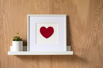 Red heart shape in a white modern frame standing on a white shelf with wooden wall on the background. Mockup fram e design for square art