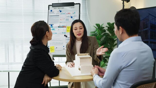 Asian businesswoman presenting an architectural model during a team meeting in a modern office, collaborating with colleagues in a professional and creative environment.