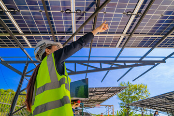 A technician in a safety helmet and reflective vest inspects a solar panel structure using a...