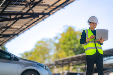 Obraz premium A female technician wearing a safety helmet and reflective vest uses a laptop while inspecting a solar carport installation. The scene features solar panels and parked cars under a clear sky.