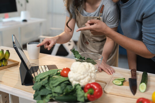 Young couple preparing a healthy meal together in a modern kitchen, following a recipe on a tablet, pointing at fresh ingredients and discussing the cooking process with joy