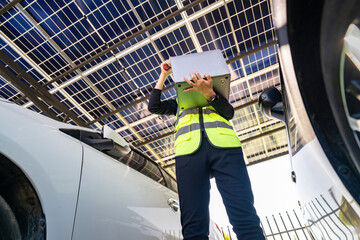 Engineer inspecting solar panels in a solar-powered carport surrounded by parked cars. Renewable...