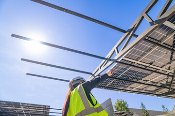 A technician in a safety helmet and reflective vest inspects a solar panel structure using a...