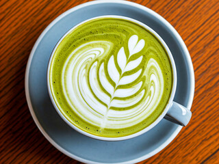 A top-down view of a vibrant green matcha latte served in a white ceramic cup on a matching blue-gray saucer. The drink features intricate latte art in the shape of a leaf, crafted with creamy white .