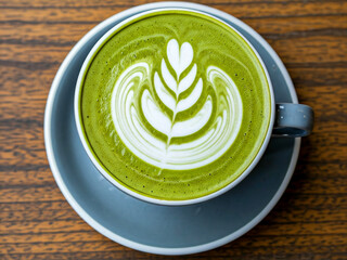 A top-down view of a vibrant green matcha latte served in a white ceramic cup on a matching blue-gray saucer. The drink features intricate latte art in the shape of a leaf, crafted with creamy white .