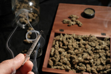 Smoking marijuana joint in a man's hand on a background of cannabis buds, grinder and jars with cones