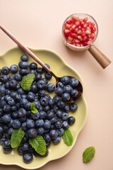 Close-up view of a green plate and copper spoon with fresh blueberries and a bowl with pomegranate, with mint leaves on beige background. Concept of healthy eating and fresh fruit