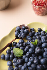 Close-up of a green plate with blueberries, a copper spoon and mint leaves with background containers with pomegranate and beige background. Healthy eating concept