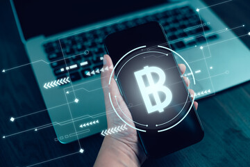 Digital currency concept featuring a glowing cryptocurrency symbol on a smartphone screen held by a hand, with futuristic virtual interface elements and a laptop in the background.