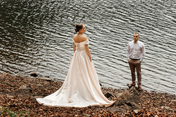 couple standing on the shore of the lake. The woman is wearing a long white wedding dress with open...