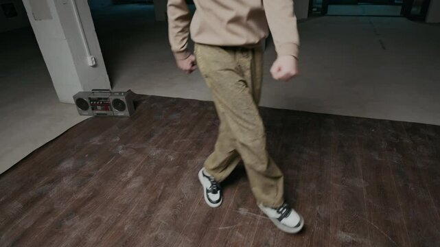 Handheld shot of talented Caucasian gen Z b-boy in streetwear performing top rock moves, improvising while breaking alone in indoor dance hall to music on boombox