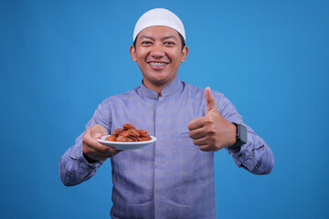 Smiling Young Asian Guy Giving Thumbs Up While Showing Dates on Plate Isolatedn on Blue Background