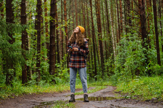 Young female hiker with backpack is using smartphone gps app enjoying hiking trail in the forest after rain, wearing yellow beanie hat and checkered shirt