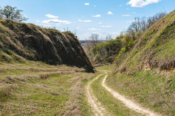 A winding dirt path cuts through steep grassy hills under a bright blue sky with scattered white clouds on a sunny day.
