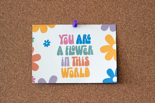 A postcard with flowers that says 'you are a flower in this world', on a corkboard, push pin