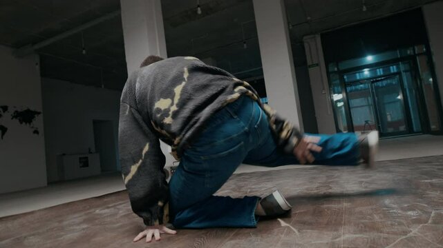 Handheld shot of talented Caucasian male zoomer in street style clothes performing down rock and power moves showing his strength and individuality while breaking to music in empty dance hall