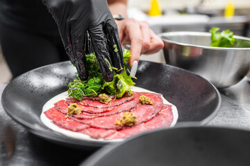 A chef's hand in black gloves carefully garnishes a plate of sliced raw meat with fresh greens and herbs, showcasing a modern culinary presentation in a professional kitchen.
