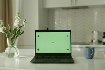 Background shot of open laptop with blank green mockup screen on counter in cozy white kitchen with minimalistic interior, copy space