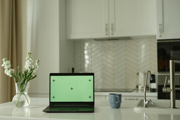 Still life shot of open laptop computer with green mockup screen placed on kitchen counter decorated with fresh flower in vase in cozy white room with scandinavian interior, copy space