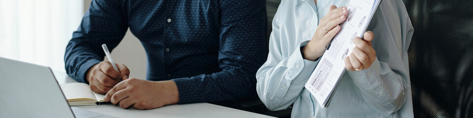Two colleagues engaged in discussing business details, one taking notes while other holding a...