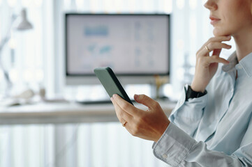 Person holding smartphone while sitting at work desk in modern office setting. Blurred computer...