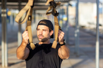 Man in black t-shirt and cap using suspension straps in an outdoor fitness session