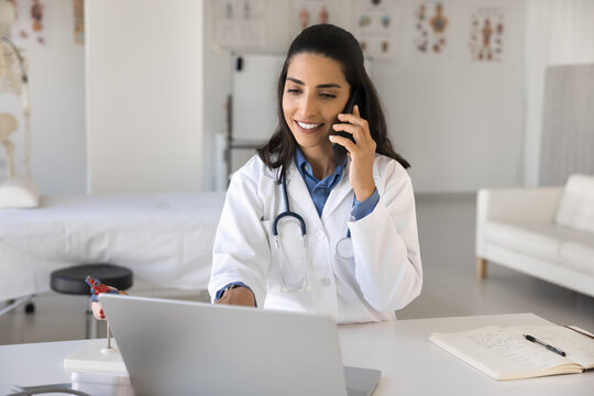 Multitasking in medicine. Smiling confident young woman doctor medical professional consult patient by phone on distance discuss treatment plan managing case history file in healthcare app on notebook