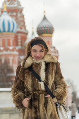 Beautiful woman in fur coat smiling in front of colorful St. Basil's Cathedral during winter in...