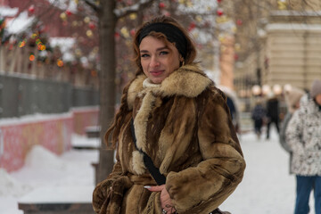 Woman in warm fur coat enjoys winter day in a snow-covered city park with festive decorations and...
